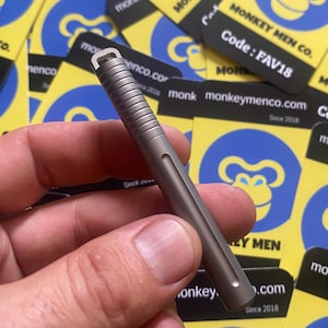 Könnte beinhalten: Ein kleiner, metallisch grauer Stift mit geriffelter Oberseite und einer gerillten Seite. Der Stift hat eine Schlaufe oben und wird vor einem Hintergrund aus gelben und blauen Karten mit einem Affenlogo und dem Text "MONKEY MEN CO." gehalten.