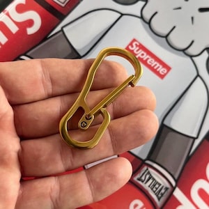 Puede incluir: Un mosquetón de color dorado, sostenido en una mano, con una puerta con resorte. El mosquetón es de diseño simple y funcional, probablemente para sujetar llaves u otros objetos pequeños. El fondo incluye un logotipo rojo y blanco.