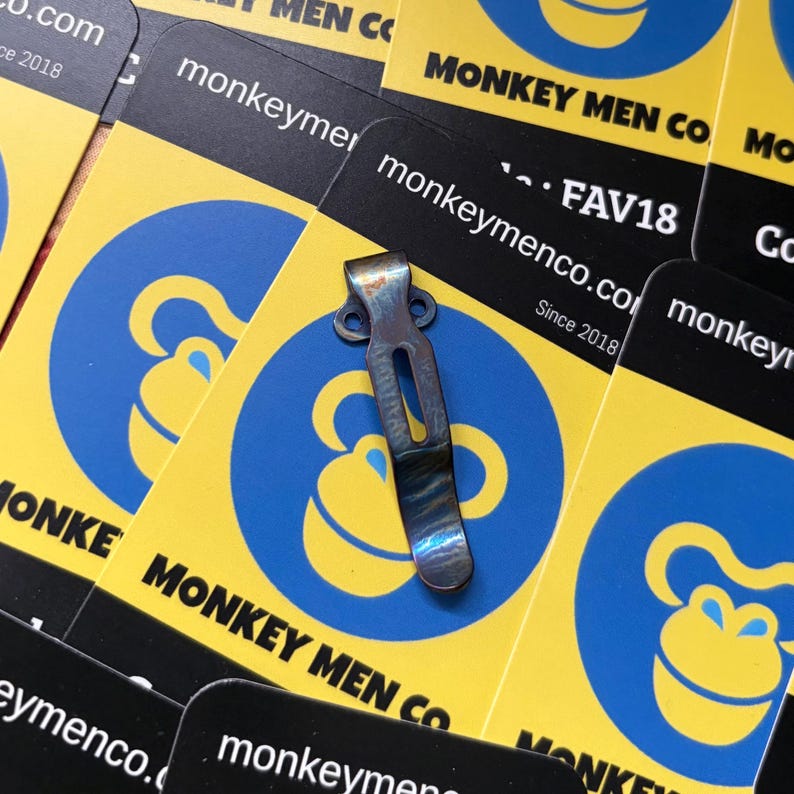 May include: A small, metallic pocket clip with a worn, iridescent finish rests on a background of yellow and black cards. The cards feature a blue monkey logo and the text "MONKEY MEN CO" and "monkeymenco.com Since 2018".