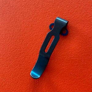 May include: A black metal pocket clip with two small holes on the top. The clip is curved and has a smooth finish.