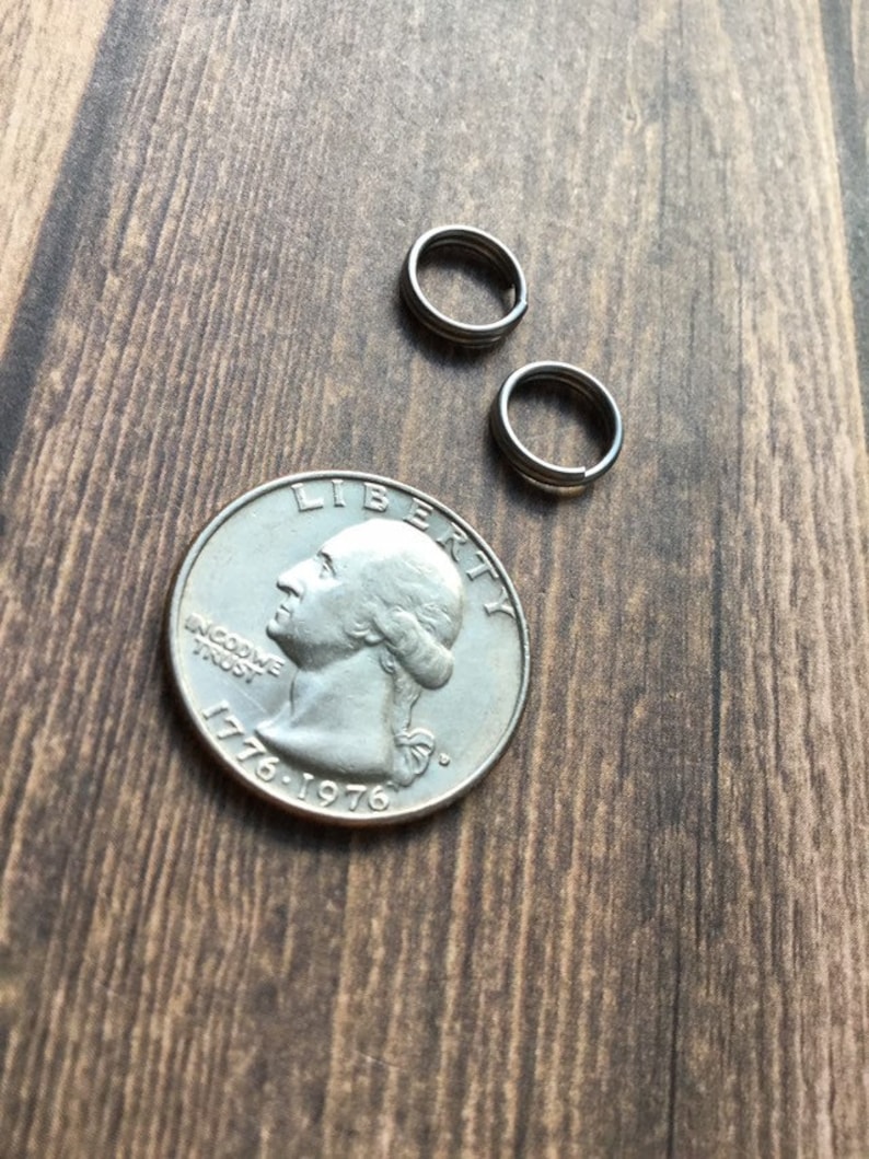 May include: Close-up of a silver quarter coin with the profile of George Washington and the years 1776-1976. Two small, silver split rings are next to the coin on a wooden surface. The rings are made of three connected loops.