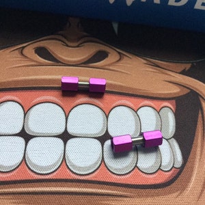 Könnte beinhalten: Zwei kleine, leuchtend pinkfarbene Metallobjekte in Hantelform, jeweils mit einem Gewindezentralstab. Die Objekte befinden sich auf einer gemusterten Oberfläche mit einem Cartoon-Gesicht und dem Wort "BENCHMADE" in Weiß auf blauem Hintergrund.