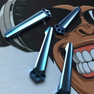 Puede incluir: Cuatro escamas de cuchillo de titanio azul con bordes redondeados y tres agujeros cada una. Las escamas est&aacute;n dispuestas en forma de abanico sobre un fondo blanco y marr&oacute;n.