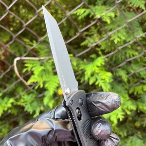 Könnte beinhalten: Ein schwarzes Klappmesser mit einer teilweise gezahnten Klinge und einem schwarzen Griff. Das Messer hat einen goldenen Akzent am Griff.
