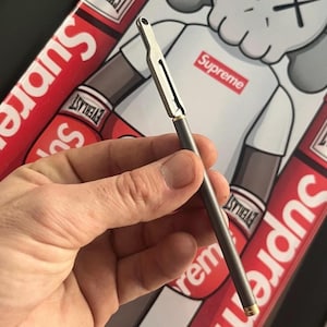 Può includere: Una penna argentata e grigia con dettagli dorati. La penna è tenuta in mano, su uno sfondo con la scritta "Supreme" in rosso e bianco.
