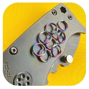 Op de afbeelding: Close-up van een grijs vouwmes met een verzameling regenboogkleurige sleutelringen. De sleutelringen zijn van metaal en hebben een cirkelvorm. Het mes heeft een getextureerd oppervlak en een gedeeltelijk zichtbaar serienummer.