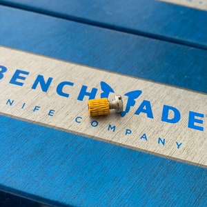 Könnte beinhalten: Nahaufnahme einer goldfarbenen Metallschraube mit geriffeltem Kopf, die auf einem blauen Kasten mit dem aufgedruckten Text "BENCHMADE KNIFE COMPANY" liegt.