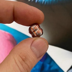 Puede incluir: Una pequeña cuenta metálica con un diseño tejido. La cuenta se sostiene entre dos dedos y presenta bandas alternas de cobre y metal plateado. La cuenta tiene una forma redondeada e irregular y refleja la luz.