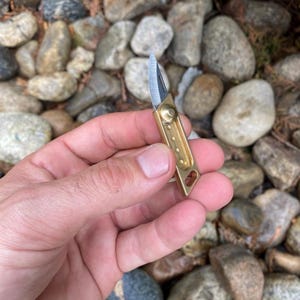 May include: A small, brass-colored folding knife with a silver blade. The knife is held in a hand against a background of rocks. The knife is compact and appears to be a keychain knife.