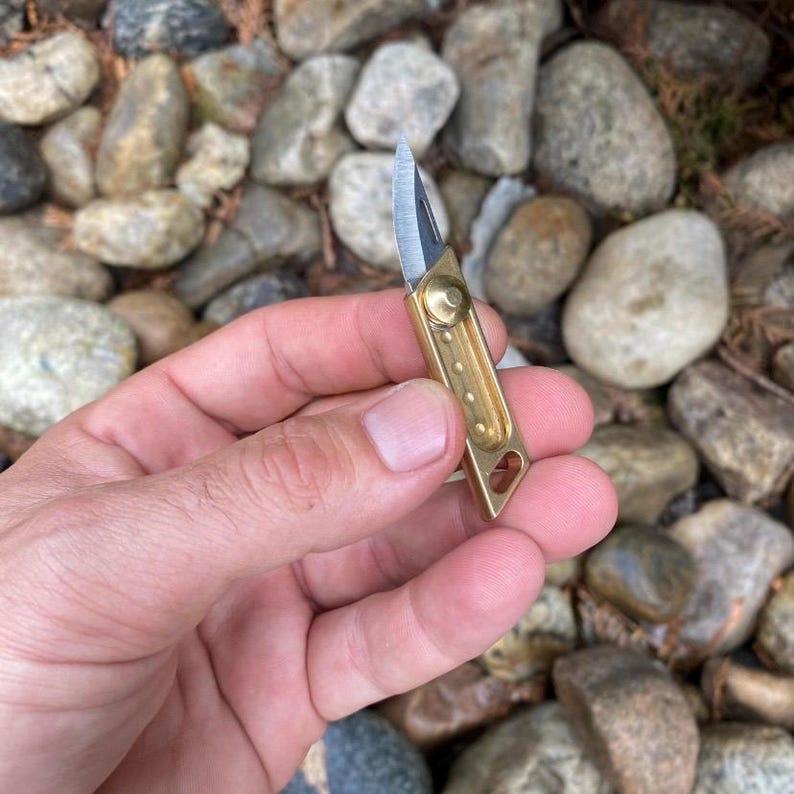 May include: A small, brass-colored folding knife with a silver blade. The knife is held in a hand against a background of rocks. The knife is compact and appears to be a keychain knife.