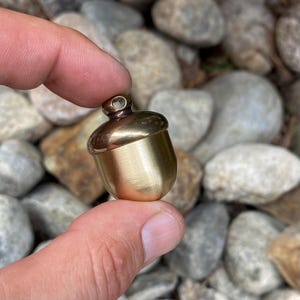 May include: A small, brass acorn pendant with a loop at the top. The acorn has a smooth, polished finish and is held between fingers against a background of rocks. The acorn's cap is a darker shade of brass than the body.