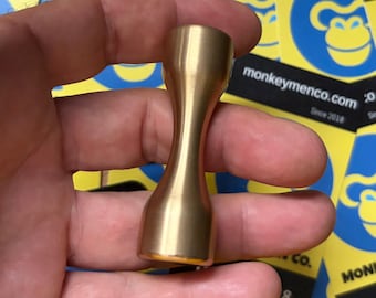 May include: A solid brass hourglass-shaped object with a smooth, metallic finish. The item features a wider base and top that taper into a narrow, cylindrical centre, creating a symmetrical, balanced form with a polished, reflective surface.