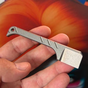 Puede incluir: Una pequeña herramienta metálica gris con una hoja plana rectangular y un mango que presenta tres ranuras diagonales y un orificio circular en el extremo. La superficie tiene un acabado mate y texturizado, con un diseño compacto y utilitario.