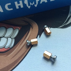 Könnte beinhalten: Drei goldfarbene Metall-Abstandshalter mit silbernen Schrauben. Die Abstandshalter sind sechseckig und haben eine glatte, glänzende Oberfläche.