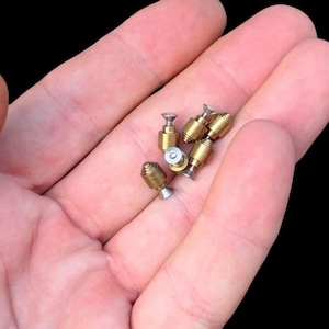 Könnte beinhalten: Eine Sammlung kleiner, messingfarbener Metallkomponenten, möglicherweise Miniaturhardware oder Modellbauteile. Die Komponenten haben eine zylindrische oder konische Form mit schraubenartigen Merkmalen. Die Artikel werden in einer Hand vor schwarzem Hintergrund gehalten.