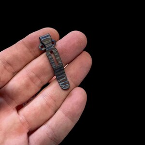 May include: A black and brown patterned metal pocket clip. The clip has a curved shape and a small hole on each end.