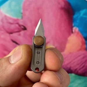 May include: A small, silver and brass folding knife with a sharp blade. The knife is held in a hand, with a pink and blue background. The blade is open, and the knife appears to be new.