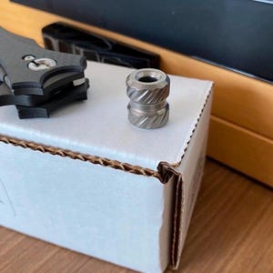 Puede incluir: Una pequeña cuenta metálica cilíndrica con un patrón diagonal texturizado. La cuenta es de color plateado y se encuentra sobre una caja de cartón blanca. Otros objetos metálicos son visibles en el fondo.