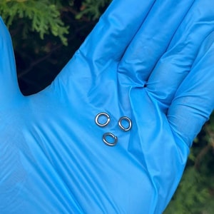 Puede incluir: Tres pequeños anillos de salto plateados se muestran sobre un fondo azul. Los anillos son circulares y parecen ser de metal. La imagen está bien iluminada, destacando los detalles de los anillos.