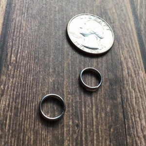 May include: Two small silver rings and a quarter coin rest on a dark wood surface. The rings are circular and appear to be made of metal. The quarter is silver and has a portrait on it.