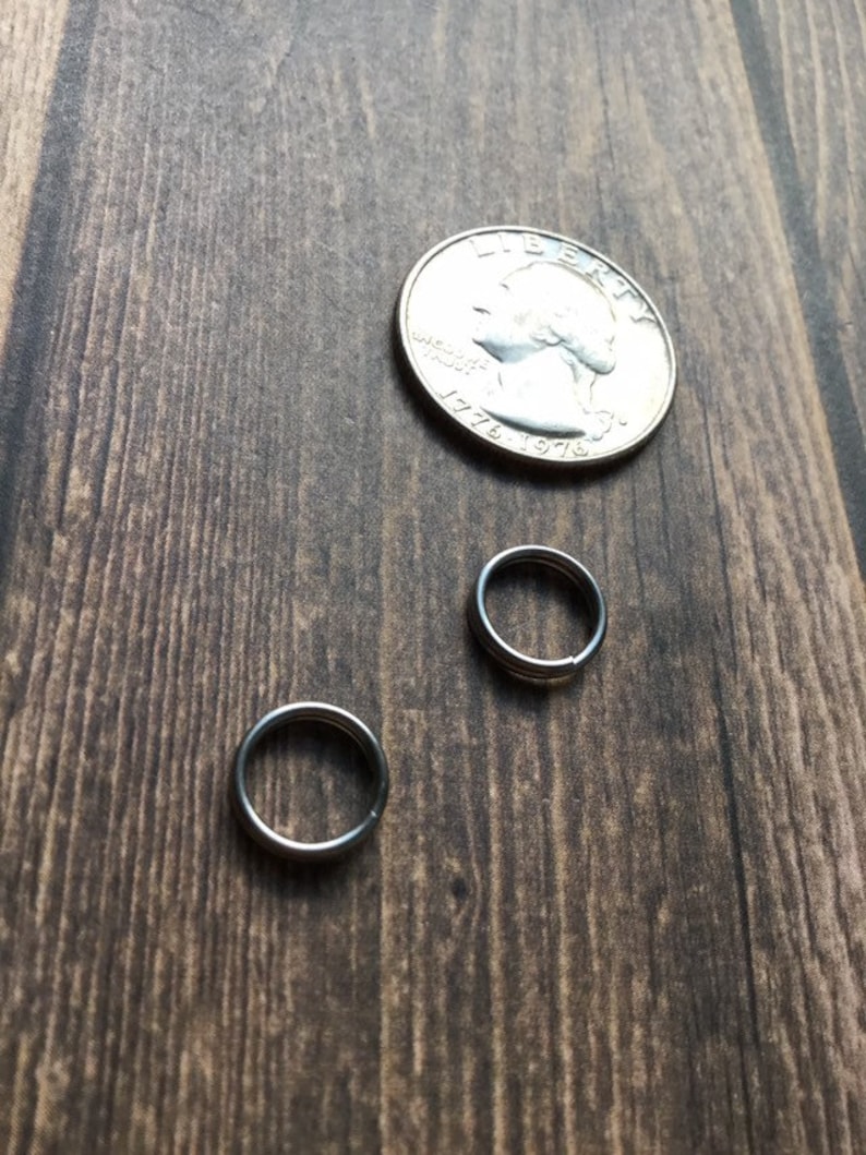 May include: Two small silver rings and a quarter coin rest on a dark wood surface. The rings are circular and appear to be made of metal. The quarter is silver and has a portrait on it.