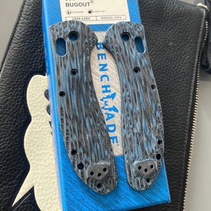 May include: Two knife handle scales with a blue and black wood grain pattern. The scales are from the Benchmade Bugout series. The packaging is blue with white text that reads "BUG OUT" and "EVERYDAY".