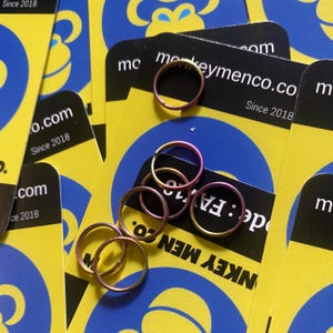 Puede incluir: Varios anillos metálicos pequeños y circulares en tonos rosa, dorado y morado se encuentran sobre un fondo de tarjetas amarillas y azules. Las tarjetas tienen el texto "monkeymenco.co Since 2018".