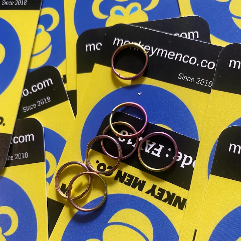May include: Several small, circular, metallic rings in shades of pink, gold, and purple are scattered on a background of yellow and blue cards. The cards have the text "monkeymenco.co Since 2018".