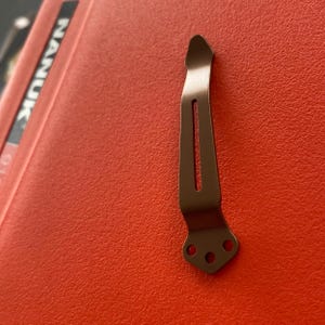 May include: A brown metal pocket clip with three holes at the bottom. The clip is designed to attach to a pocket or belt.