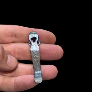 May include: A blue and copper-toned titanium pocket clip with a textured finish. The clip has a unique shape with a small hole at the top.