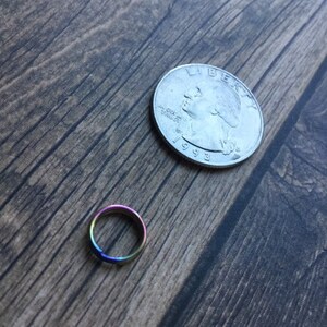 May include: A small, iridescent ring with a rainbow finish, next to a U.S. quarter coin on a wooden surface. The ring's colors shift from purple to blue, green, and yellow. The quarter is dated 1993.
