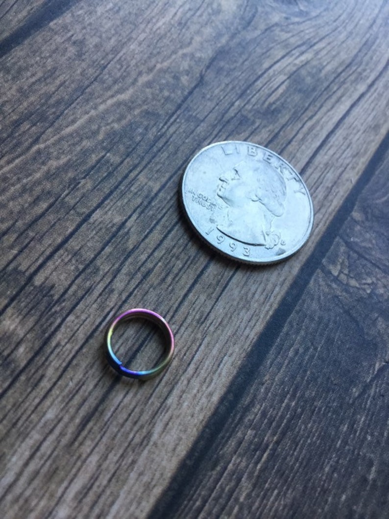 May include: A small, iridescent ring with a rainbow finish, next to a U.S. quarter coin on a wooden surface. The ring's colors shift from purple to blue, green, and yellow. The quarter is dated 1993.
