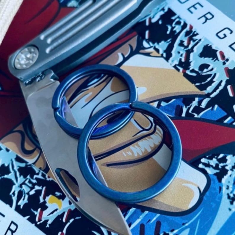 May include: Close-up of a multi-tool knife and two blue key rings. The knife is silver with a small light. The key rings are circular and have a blue iridescent finish. The background has a colorful graphic design.