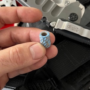 K&ouml;nnte beinhalten: Eine kleine, zylindrische Perle mit einem hellblauen und silbernen Design. Die Perle hat eine strukturierte Oberfl&auml;che mit einem Wabenmuster. Die Perle wird in der Hand einer Person gehalten, mit einem Messer und anderen Werkzeugen im Hintergrund.