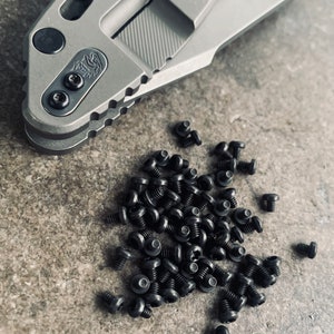 May include: A close-up of a pile of black screws on a gray textured surface. The screws are small and have a flat head. There is a gray metal object in the background.