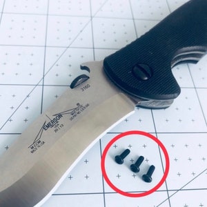 May include: Close-up of three black screws on a white surface with a black and silver knife in the background. The knife has the text "EMERSON USA 2013 M-COM MADE IN USA WAVE FEATURE US PAT NO 5878500 3160" on the blade.