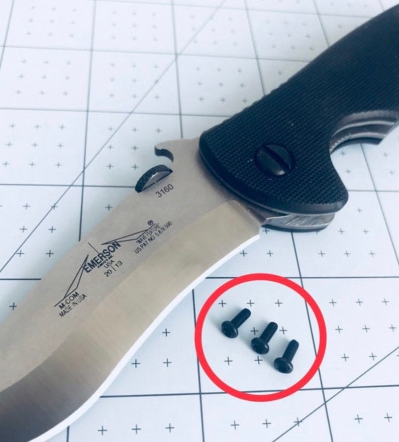 May include: Close-up of three black screws on a white surface with a black and silver knife in the background. The knife has the text "EMERSON USA 2013 M-COM MADE IN USA WAVE FEATURE US PAT NO 5878500 3160" on the blade.