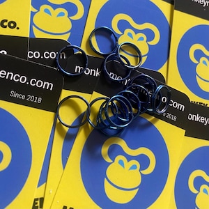 Puede incluir: Un conjunto de pequeños anillos metálicos azules sobre un fondo de tarjetas amarillas con un logotipo de mono azul. Las tarjetas también presentan el texto "enco.com Since 2018". Los anillos son circulares.