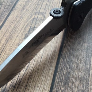 May include: A silver folding knife with a black handle. The blade has the text "EMERSON KNIVES" and "MADE IN USA" on it.