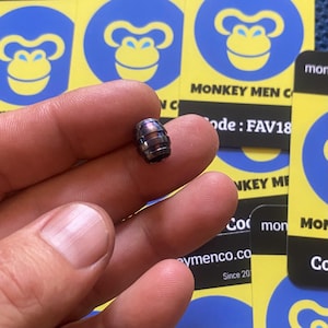 Puede incluir: Una pequeña cuenta en forma de barril con un acabado metálico, sostenida entre los dedos. La cuenta tiene un color oscuro e iridiscente con bandas azules, moradas y bronce. El fondo incluye tarjetas amarillas con un logotipo de mono y el texto "MONKEY MEN C" y "ymenco.co Since 201".