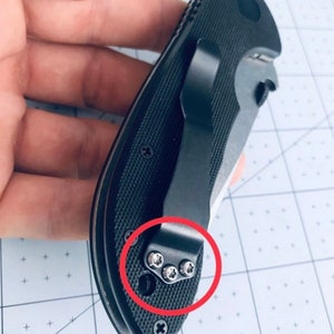 May include: A black pocket knife with a clip and a textured handle. The clip is attached to the handle with four silver screws.