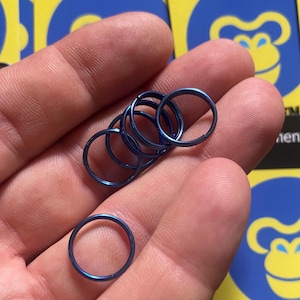 Puede incluir: Un conjunto de anillos metálicos azules se muestra en una mano. Los anillos son de un azul metálico brillante. Los anillos son circulares y parecen estar hechos de un material duradero. El fondo está borroso, con un logotipo visible.
