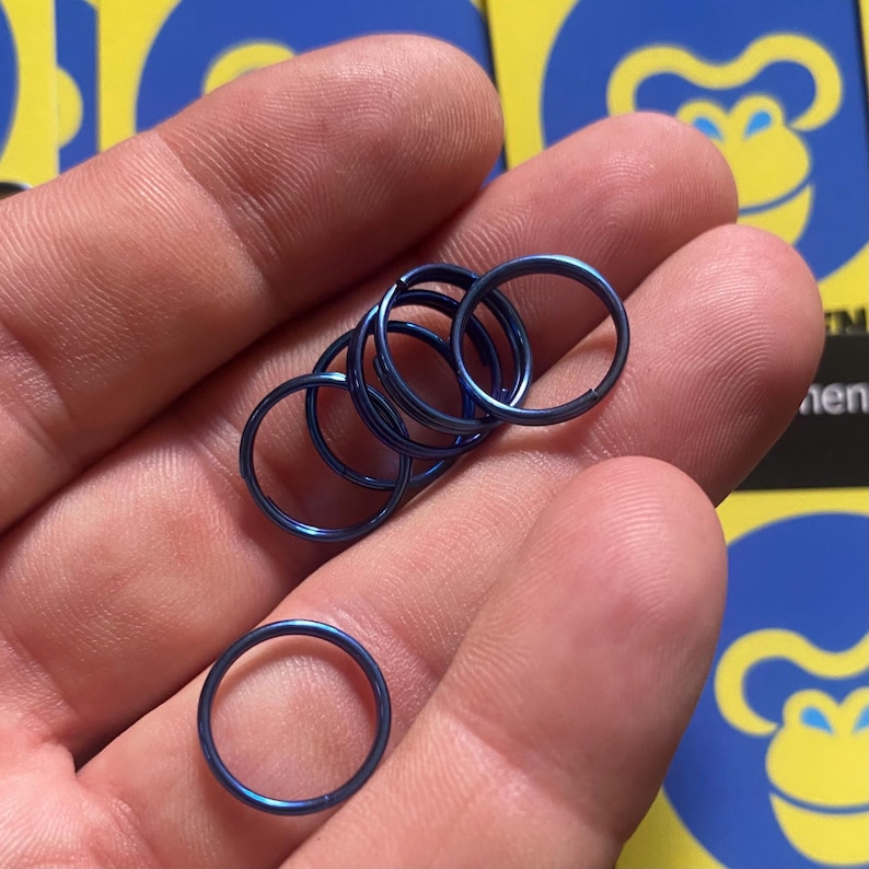 May include: A collection of blue metal rings, varying in size, are held in a hand. The rings are a vibrant, metallic blue color. The rings are circular and appear to be made of a durable material. The background is blurred, with a logo visible.