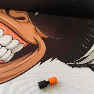 Könnte beinhalten: Eine schwarze und orangefarbene Metallschraube mit einem gerändelten Kopf. Die Schraube befindet sich auf einer weißen Oberfläche mit einem schwarzen und braunen Comic-Affen im Hintergrund.