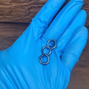 Puede incluir: Tres pequeños anillos de metal gris oscuro conectados en cadena. Los anillos son circulares y parecen estar hechos de metal macizo. Los anillos se sostienen en una mano enguantada de azul.
