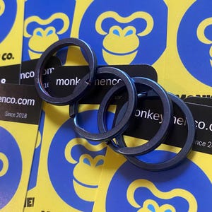 May include: Four dark gray key rings are displayed on a background of yellow and blue cards. The cards feature a monkey logo and the text "monkey nence co." and "enco.com Since 2018".