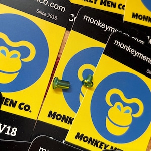 Peut inclure: Gros plan sur des fixations métalliques vertes, possiblement des vis ou des rivets, sur un fond de cartes jaunes et noires. Les cartes présentent un cercle bleu avec un logo de singe jaune et le texte "monkeymen.com Since 2018".
