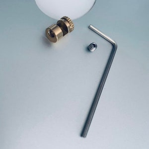 Könnte beinhalten: Ein Messing-Kameragurt-Verbinder mit einem kleinen Inbusschlüssel und einer Ersatzschraube. Der Verbinder hat ein Gewindeloch und einen kleinen Knopf zum Einstellen der Gurtlänge.