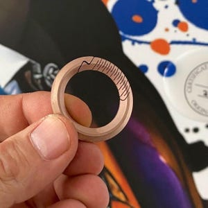 Puede incluir: Un anillo de madera marrón claro con un diseño decorativo de líneas negras. El anillo se sostiene en una mano, mostrando sus intrincados detalles y artesanía. El diseño del anillo presenta una serie de líneas paralelas y una línea curva.
