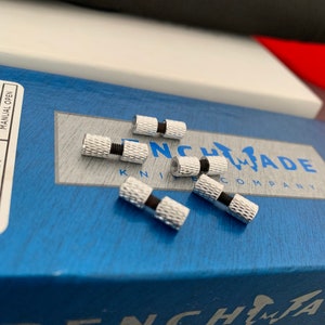 Könnte beinhalten: Nahaufnahme von sechs silbernen Metallschrauben mit schwarzen Gummidichtungen. Die Schrauben befinden sich auf einer blauen Box mit dem aufgedruckten Text "BENCHMADE KNIFE COMPANY".
